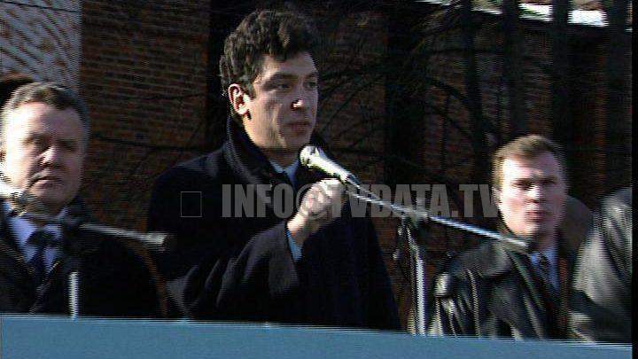 Photograph of Boris Nemtsov, a handsome Russian politician, speaking passionately on a stage during a political rally on a beautiful autumn day in Moscow, Russia. He is dressed in a suit, gesturing expressively to an unseen crowd. The backdrop features golden autumn trees and historic Moscow architecture under a clear blue sky